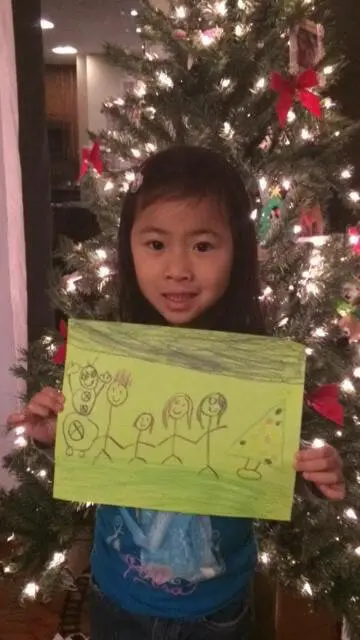 Child posing with a holiday artwork in front of a Christmas tree for the Alexandria Children's Dentistry Holiday Art Raffle