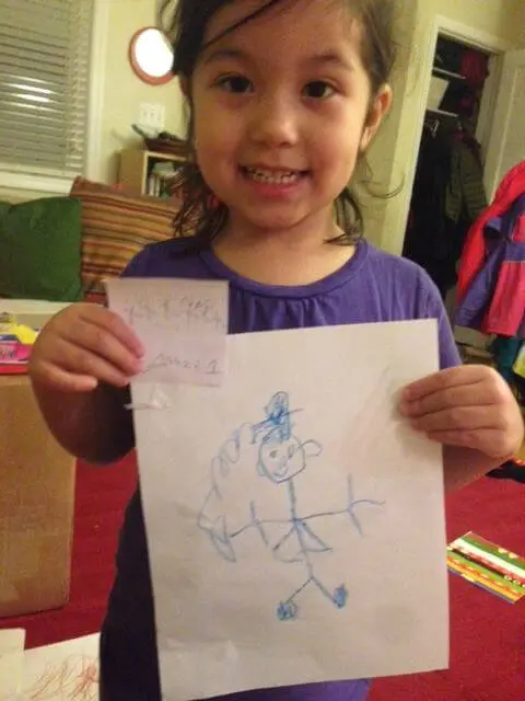 Child smiling while showing a hand-drawn winter artwork for the Alexandria Children's Dentistry Holiday Art Raffle