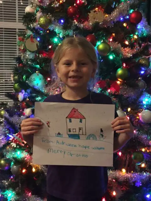 Child holding holiday artwork beside a Christmas tree for the Alexandria Children's Dentistry Holiday Art Raffle