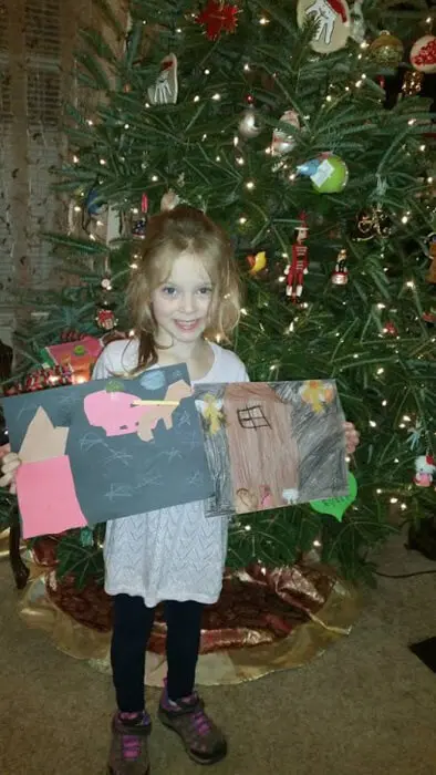 Child posing with holiday artwork beside a decorated Christmas tree for the Alexandria Children's Dentistry Holiday Art Raffle