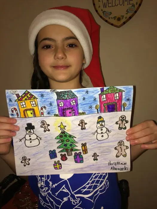 Child in a Santa hat holding a colorful Christmas village drawing for the Alexandria Children's Dentistry Holiday Art Raffle