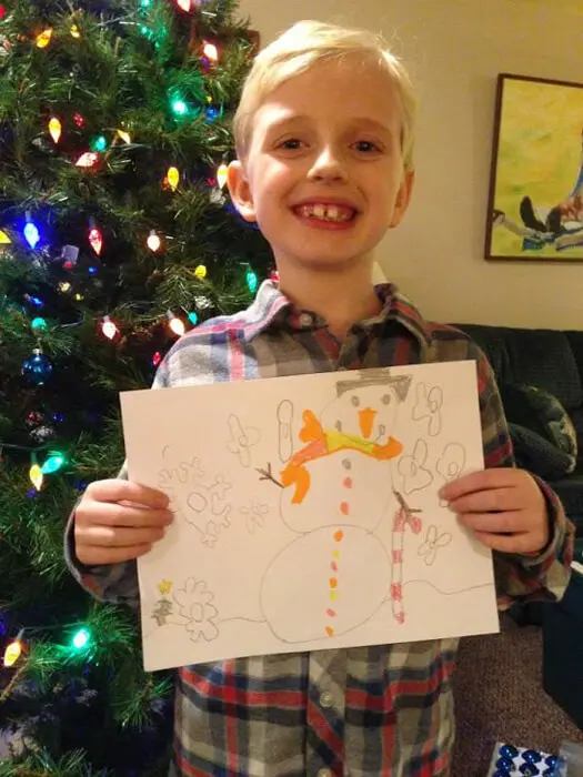Child smiling while holding a snowman drawing for the Alexandria Children's Dentistry Holiday Art Raffle