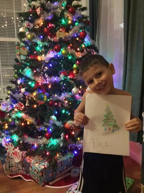 Child holding a Christmas tree drawing beside a decorated tree for the Alexandria Children's Dentistry Holiday Art Raffle